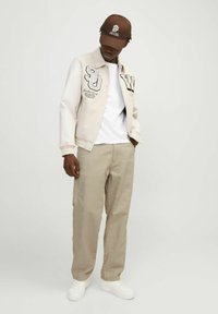Chaqueta varsity blanca y beige con letras y gráficos, llevada sobre una camiseta blanca, combinada con pantalones beige y zapatillas blancas. Gorra marrón.