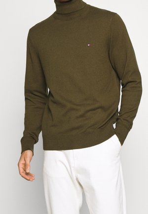 Man draagt een olijfgroene coltrui met een klein logo op de borst en witte broek, staat met één hand in de zak.