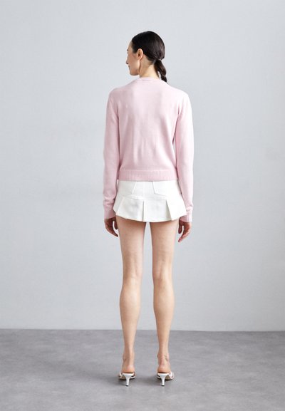 Light pink knitted sweater with ribbed cuffs, paired with a white pleated mini skirt. The outfit is completed with white heels.