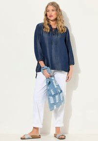 Blusa in denim navy con maniche a tre quarti e scollatura a V con lacci; abbinata a pantaloni bianchi leggeri e giacca in denim strappata. Sandali argentati.