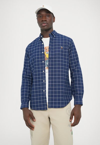 Temno modra karo srajca z belo mrežasto vzorcem, ovratnikom z gumbi in majhnim logotipom na prsih. Nošena preko grafične majice.