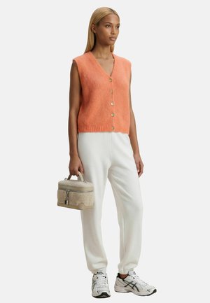 Femme debout face à l’avant, portant un gilet orange sans manches boutonné, un pantalon blanc, des baskets blanches et tenant un sac à main beige texturé.