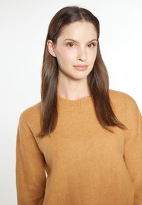 Pulover de culoare muștar, cu un guler rotund tricotat, realizat dintr-un material moale din tricot. Modelul are părul neted, machiaj minimal și un fundal neutru.