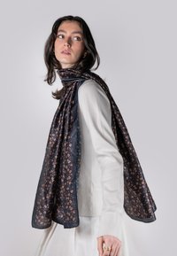 Jūras zila ziedu šalle, izgatavota no viegla auduma, ar mazām oranžām ziedu kompozīcijām, apsieta ap modeļa kaklu, izceļot mīksto tekstūru un rakstu.