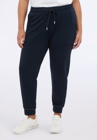 Granatowe joggery wykonane z miękkiego materiału, z elastycznym paskiem w talii i ściągaczem, bocznymi kieszeniami oraz dopasowanymi mankietami. Ubrane z białymi sneakerami.