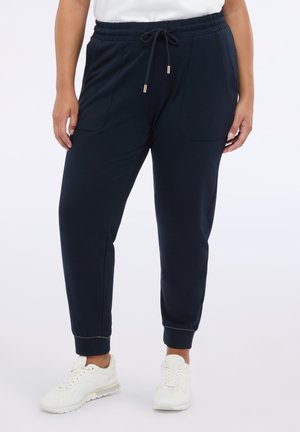 Pantalons de jogger bleu marine en tissu doux, dotés d'une taille élastique avec cordon de serrage, de poches latérales et de poignets ajustés. Portés avec des baskets blanches.