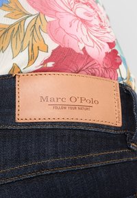 Los jeans de mezclilla de un azul oscuro presentan un parche de cuero marrón con el texto en relieve "Marc O'Polo" y "SIGUE TU NATURALEZA," destacando los detalles de costura.