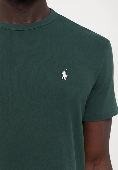 T-shirt court à manches courtes vert foncé en tissu côtelé, arborant un petit logo brodé blanc d'un joueur de polo sur la poitrine gauche.