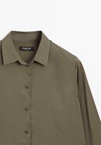 Chemise olive verte à boutons, fabriquée en tissu doux, avec un col classique et des manches longues. Étiquette visible au niveau du col.