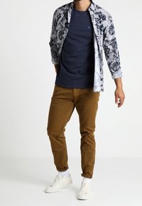 Men's outfit featuring a floral patterned white long-sleeve shirt, navy t-shirt, brown pants, and white sneakers. Cotton materials visible.