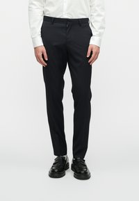 Calças de fato azul marinho com uma textura suave, ajuste slim, dois bolsos laterais e passantes para cinto, combinadas com sapatos pretos sem atacadores.