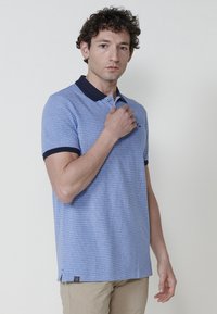 Gaiši zils polo krekls ar teksturētu audumu, tumši zila apkakle un piedurknes, neliela logo uz krūšu daļas un sānu šķirums apakšmalā.