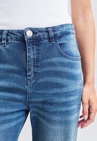 Jeans en denim bleu au design classique, avec une fermeture à bouton, des poches avant et une texture décolorée. Légèrement fuselés au niveau de la cheville.