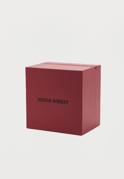 Κόκκινο ορθογώνιο κουτί με μαύρο κείμενο "HUGO KREIT" στην πρόσοψη, απλό φόντο, εμφανείς διπλωμένες άκρες στην κορυφή.