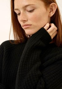 Schwarzer Rippenpullover mit hohem Kragen und langen Ärmeln, der eine weiche Textur und eine lockere Passform bietet, die teilweise die Hände bedeckt.