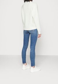 Suéter blanco de textura suave combinado con jeans ajustados azules. El modelo lleva zapatillas blancas. Costuras visibles y los jeans tienen un ligero desvanecimiento.