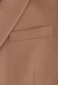 Tissu marron d'un vêtement sur mesure, avec une poche à rabat plate et un revers net ; texture lisse, lignes épurées, et design structuré.