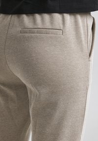 Pantalon beige texturé avec une coupe slim, une taille élastique et une poche discrète à l'arrière. Le tissu présente un motif tissé.
