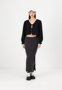 Body  cropped nera con dettaglio a fiocco, abbinata a una gonna grigia scura con spacco alto. La modella indossa un cappellino rosa chiaro e scarpe nere chunky.