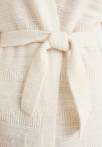Beige stickad cardigan med horisontella ränder och ett knytbälte i midjan, tillverkad av mjukt, strukturerat material.