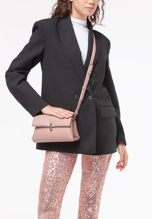 Borsa a tracolla in morbido cuoio rosa pallido con pattina pieghevole e dettagli in oro, indossata su un blazer nero e pantaloni di paillettes.