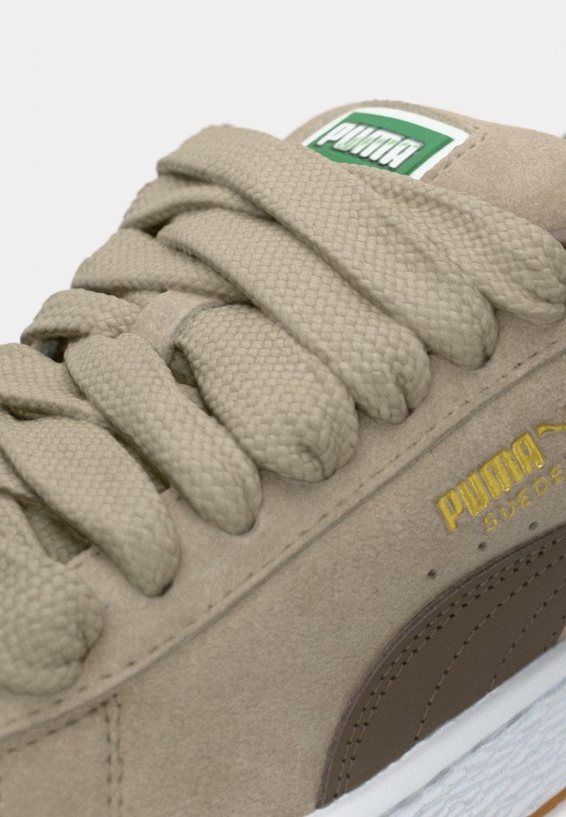 Zapatillas de ante beige con cordones texturizados y un acento de cuero marrón. Presenta una marca en verde y blanco, y una suela de goma blanca.