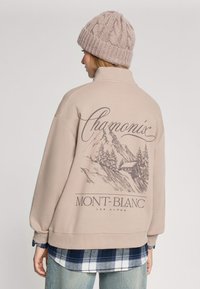 Beżowy sweter z wysokim kołnierzem, z grafiką gór oraz napisem "Chamonix Mont-Blanc". Nosi się go na niebieskiej koszuli w kratę.