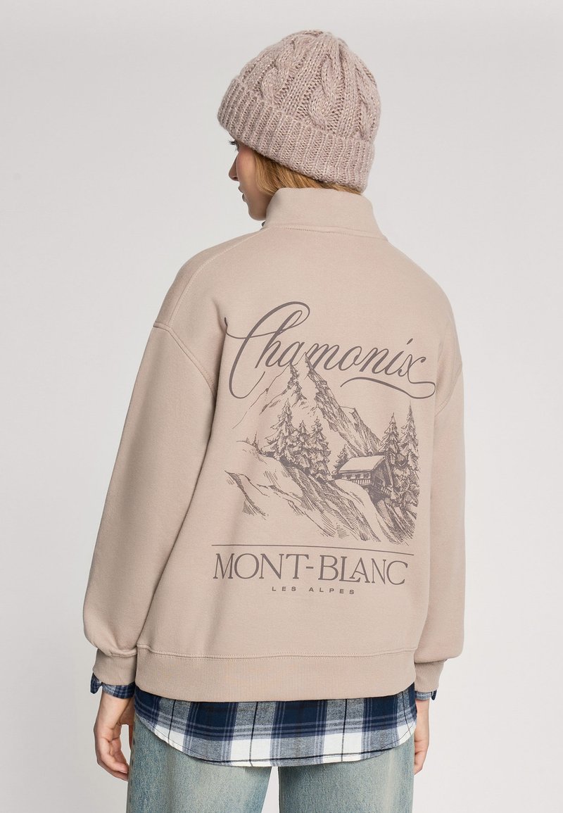 Beżowy sweter z wysokim kołnierzem, z grafiką gór oraz napisem "Chamonix Mont-Blanc". Nosi się go na niebieskiej koszuli w kratę.