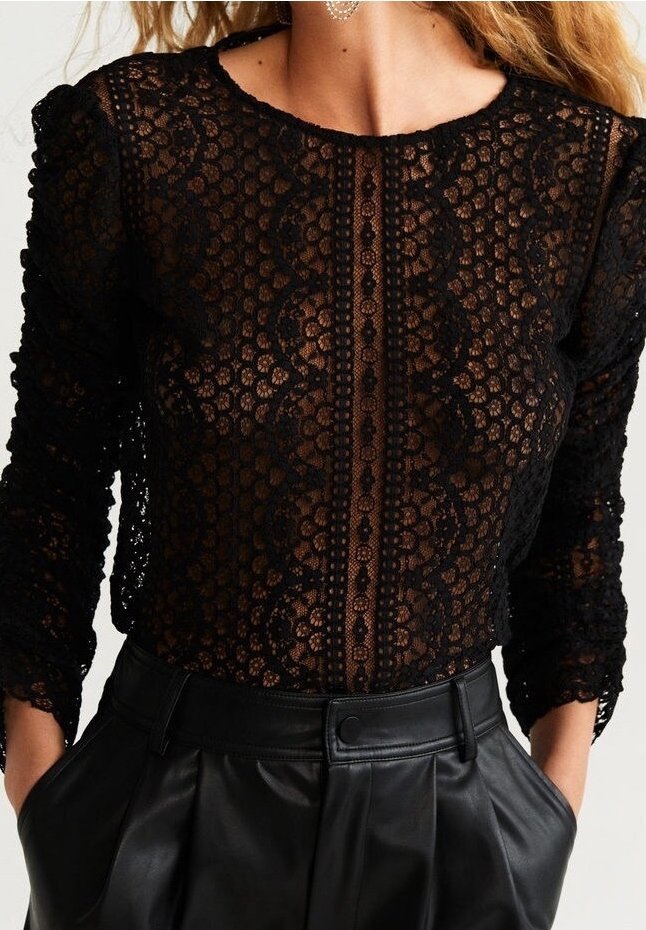 Femme portant un haut en dentelle noir à manches longues avec motifs floraux et un pantalon en cuir noir taille haute, les mains dans les poches.