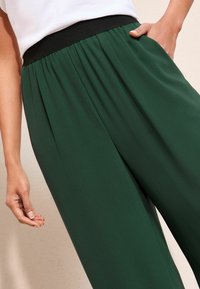 Groene wijde pantalon met plooien, voorzien van een zwarte elastische tailleband en zijzakken. Zachte stof met een gladde textuur.