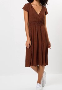 Brą dress w brązowym wzorze w zygzaki, krótkie rękawy, dekolt w serek oraz smockowany pas, zestawione z białymi trampkami. Długość sięga poniżej kolan.
