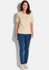 Lächelnde Frau mit braunem Haar, die ein strukturiertes beiges Kurzarmoberteil, blaue Jeans und weiße Sneakers trägt, vor einem weißen Hintergrund steht.