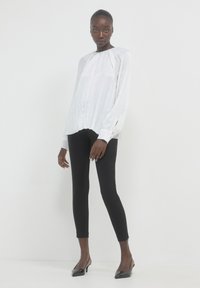 Blusa bianca plissettata con maniche lunghe, scollatura rotonda e spacco laterale, abbinata a leggings neri e scarpe col tacco nere.