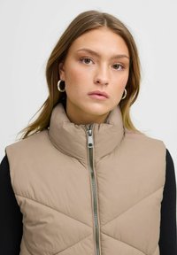 Beige pufferväst med hög krage, med en framsidan dragkedja och diamantquiltad struktur. Bärs över en svart långärmad tröja.