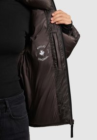 Pruun isoleeritud jakk, millel on sile ja läikiv sisevooder Khujo logoga ning tõmblukuga avaus. Kuvatud tekstuuriga väliskangas.