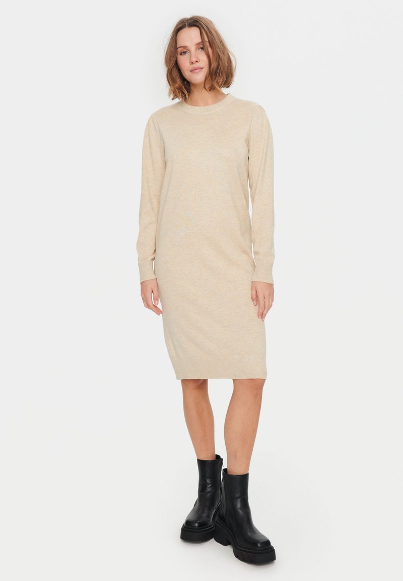 Kvinna som bär en beige stickad klänning med lång ärm och svarta klumpiga ankelstövlar, stående mot en helt vit bakgrund.