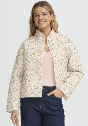 Ženska v kremno prešiti jakni z majhnim cvetličnim vzorcem, oblečena v svetlo roza majico in temno modre kavbojke, nasmejana na enobarvni podlagi.