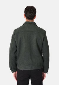 Giacca in misto lana verde con taglio dritto, ampio colletto e polsini con bottoni. Dettagli cuciti sul retro, abbinata a pantaloni scuri.