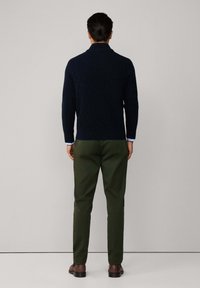 Maglione navy a lavorazione cable-knit con colletto alto, abbinato a pantaloni verde oliva. L'outfit è completato da scarpe marroni scure e sottili polsini di camicia blu.