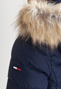 Granatowa kurtka puchowa z kapturem z sztucznego futra, z prostokątną naszywką z logo w kolorach czerwonym, białym i niebieskim na rękawie.