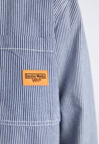 Blue and white striped workwear shirt with a chest pocket; features a rectangular orange "Service Works" label with black text.