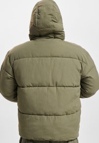 Padded olivgrüne Jacke mit Kapuze, ausgestattet mit horizontalen gesteppte Paneelen und einer weichen, matten Textur. Die Rückansicht zeigt verstellbare Details.