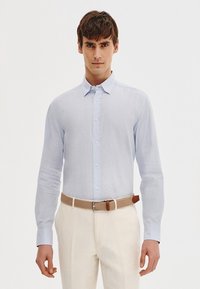 Jasnoniebieska koszula z długim rękawem i kołnierzykiem zapinanym na guziki, w połączeniu z kremowymi spodniami i brązowym plecionym paskiem z akcentem skórzanym.