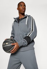 Sivý teplákový komplet s bielymi a čiernymi pruhmi, zipsom na kapucni a čiernym basketbalom. Obsahuje elastické manžety a logo na hrudi.
