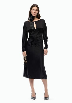 Mujer con un vestido largo de satén negro con cintura fruncida, escote con abertura, accesorio floral en el cuello, sosteniendo un pequeño bolso con cadena dorada.