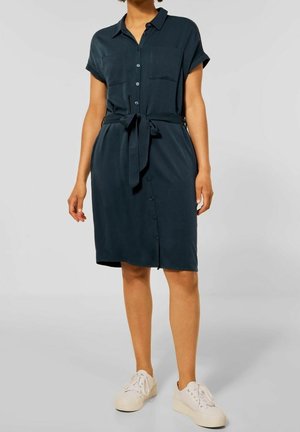 Robe chemise bleu sarcelle foncé à manches courtes, avec deux poches avant, une taille nouée et une coupe droite, portée avec des baskets blanches.