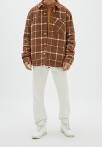 Brązowa marynarka w kratę z białymi liniami, z zapięciem na guziki i pojedynczą kieszenią na piersi, noszona na musztardowym swetrze i białych spodniach.