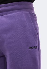 Κοντινή λήψη μοβ φόρμας με ελαστική μέση, πλαϊνή τσέπη και μαύρο κεντημένο λογότυπο "BORG" κοντά στην τσέπη.