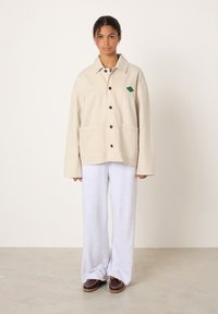 Beige Baumwolljacke mit Kragen, Knopfleiste, zwei Vordertaschen und einem grünen Logopatch; kombiniert mit grauen Jogginghosen und braunen Schuhen.