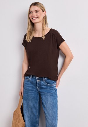 Femme souriante aux cheveux blonds portant un haut marron foncé à manches courtes et un jean bleu, tenant un sac fourre-tout en daim beige.